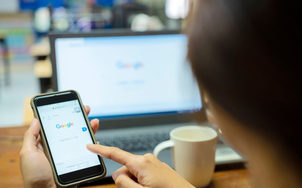 Person using Google search on a smartphone with a laptop and coffee cup in the background.