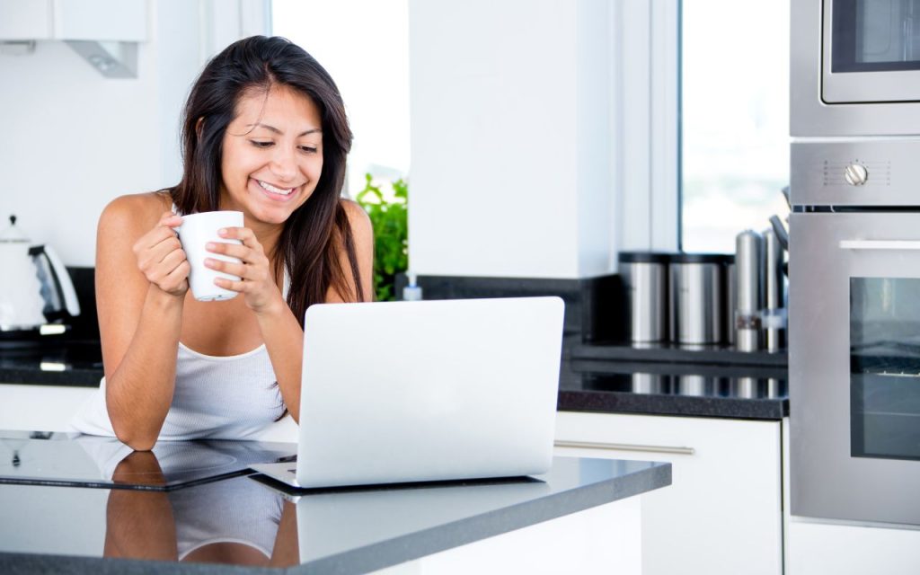 A woman smiles at a laptop screen while holding a mug, standing in a modern kitchen with white cabinets and stainless steel appliances.