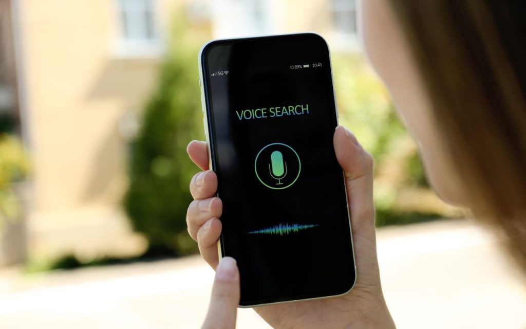 Person holding a smartphone with a voice search screen, showing a microphone icon and soundwave graphic.
