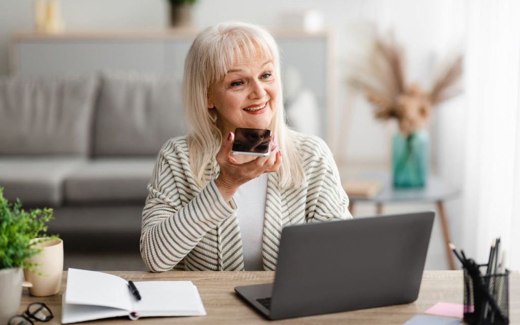 Elderly woman holding a smartphone near her mouth, sitting at a desk with a laptop, notebook, and potted plant, smiling.