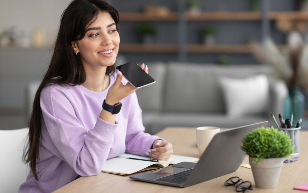 Woman in a purple sweater holds a smartphone near her mouth, sitting at a table with a laptop and potted plant, smiling in a modern room.