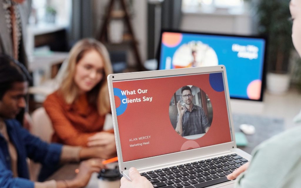 A person holds a laptop displaying a presentation slide titled "What Our Clients Say" in an office with three people working at a table in the background.