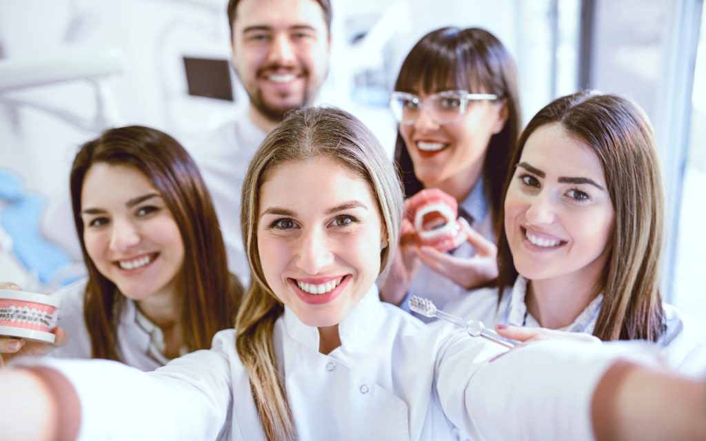 Social media isn't dead - dental clinics should adapt not abandon. Five young adults in dental uniforms smile at the camera, with two holding dental models and one holding a toothbrush, suggesting a dental or orthodontic setting.